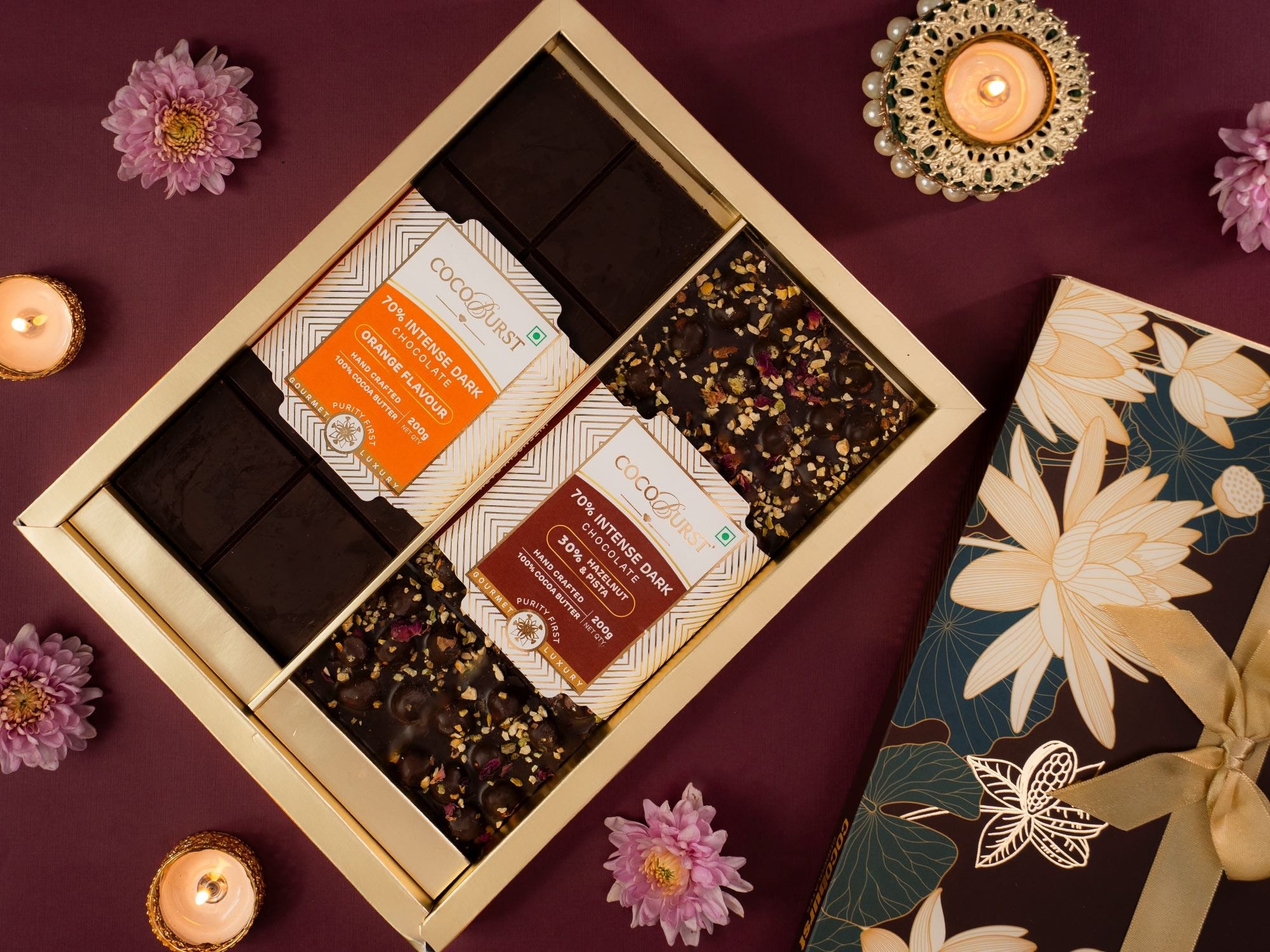 Box of chocolates with floral packaging on a purple surface with candles and flowers.